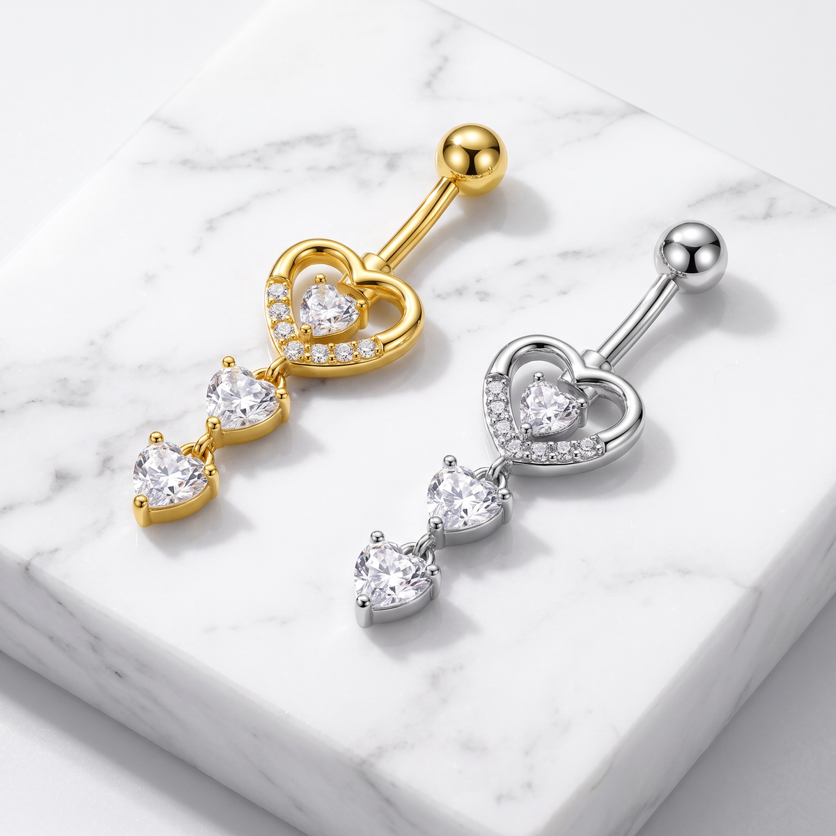 Two heart-shaped dangling belly button rings, one in yellow gold and one in silver, both featuring clear heart-shaped gemstones, displayed on a white marble block.