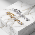 Two triple-stone belly button rings displayed on a white marble block. One ring is gold with marquise-cut clear stones, and the other is silver with marquise-cut clear stones.