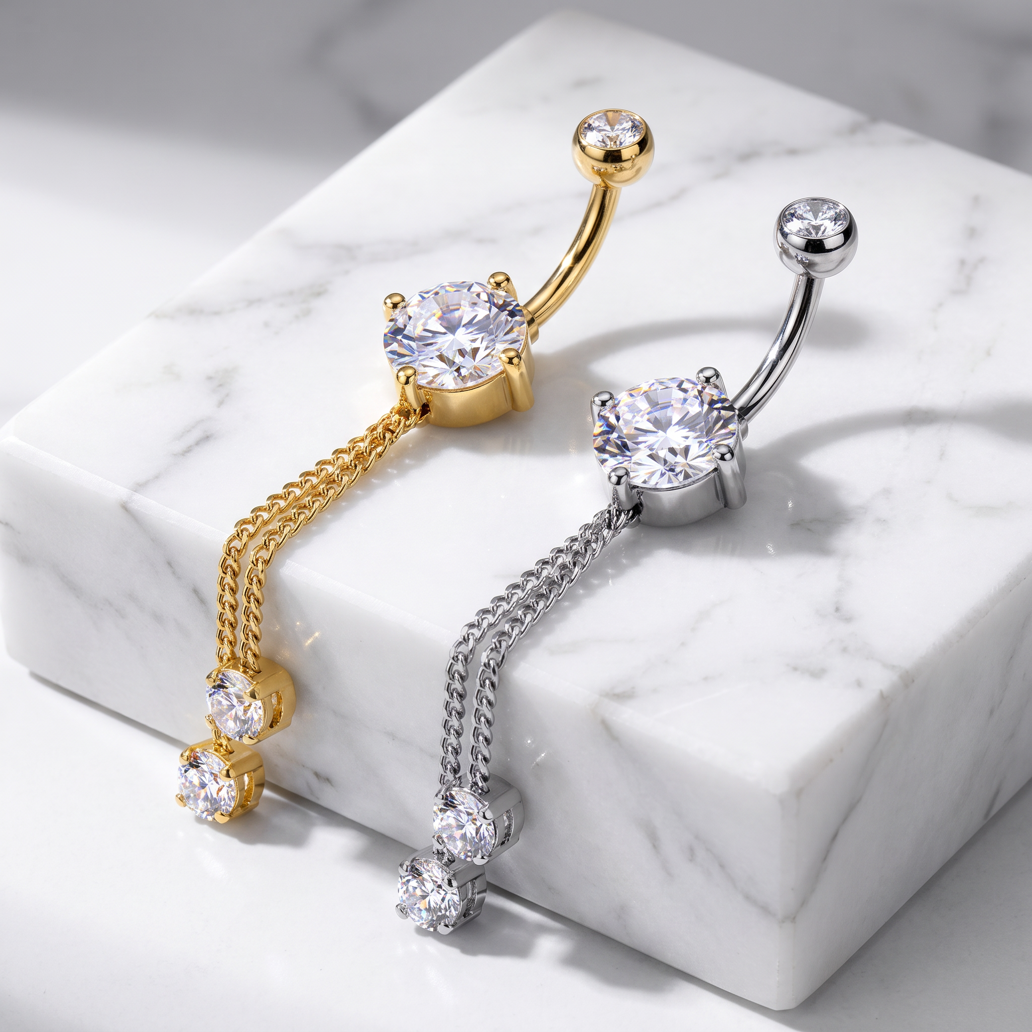 Two belly button rings with dangling chain and gemstone designs, shown on a white marble block. One ring is gold-toned and the other is silver-toned, both featuring clear cubic zirconia stones.