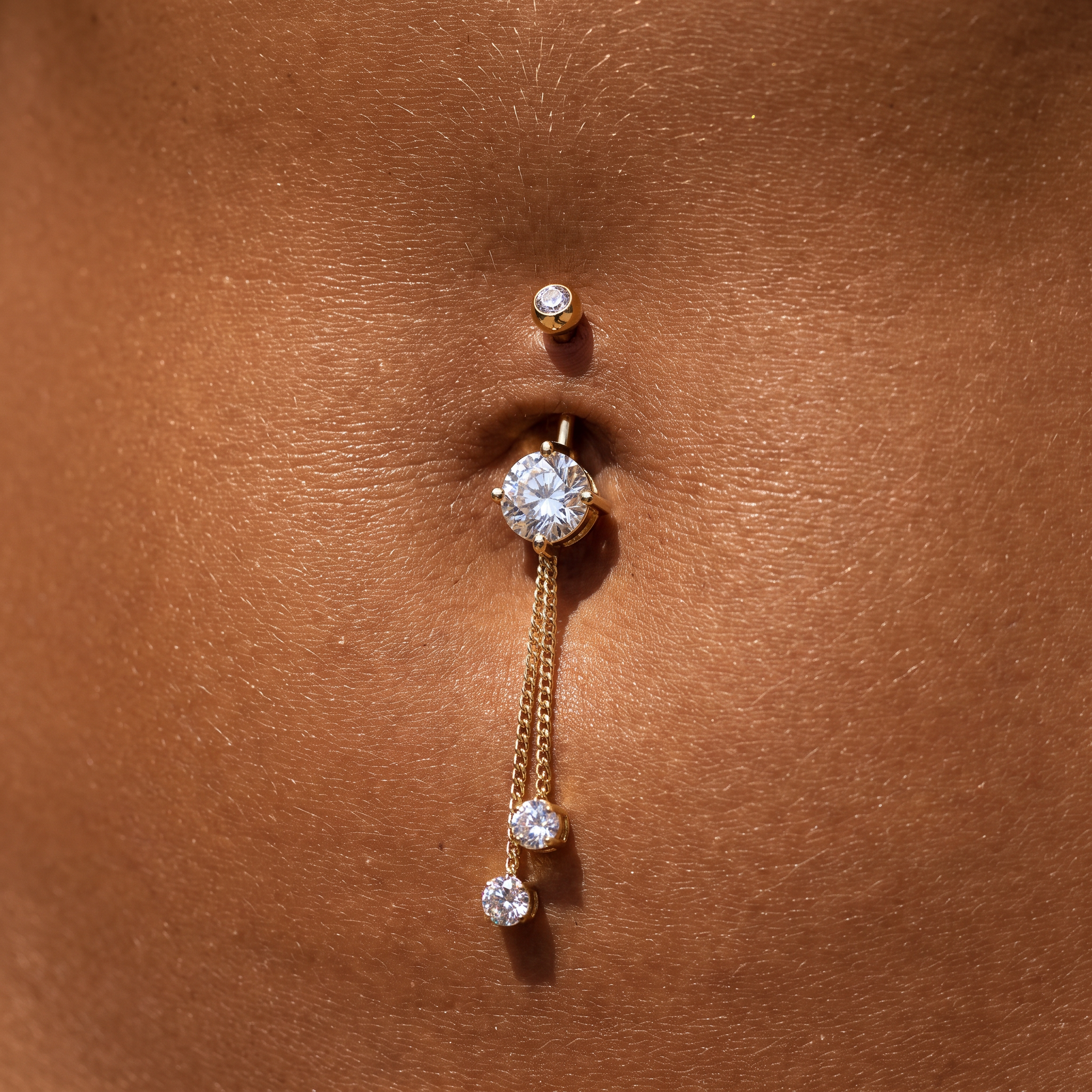 Gold belly button ring with a large round clear gemstone and two dangling chain drops, shown worn on a person's navel.