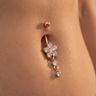 A close-up of a gold navel piercing with a dangling flower-shaped charm set with clear crystals, shown on a model's lower abdomen.