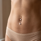 A close-up of a person's lower torso wearing a rose gold belly button ring with clear teardrop and round gemstones, paired with beige underwear.