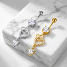Two belly button rings with dangling heart designs, one in silver and one in gold, displayed on a white marble block.