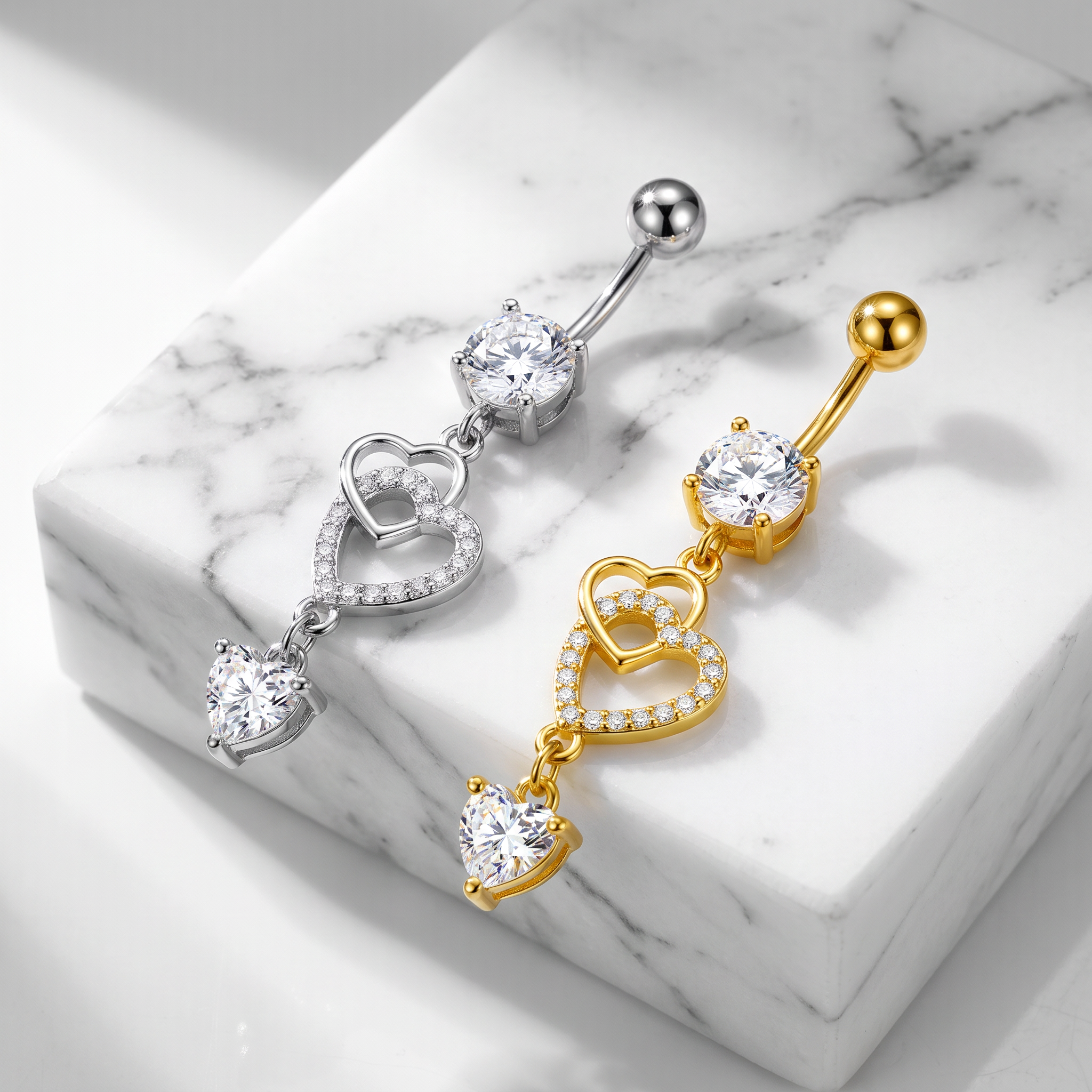 Two belly button rings with dangling heart designs, one in silver and one in gold, displayed on a white marble block.