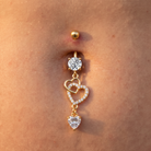 Gold belly button ring with a round clear gemstone, interlocking heart charms outlined with small clear stones, and a dangling heart charm, shown worn on a person's abdomen.