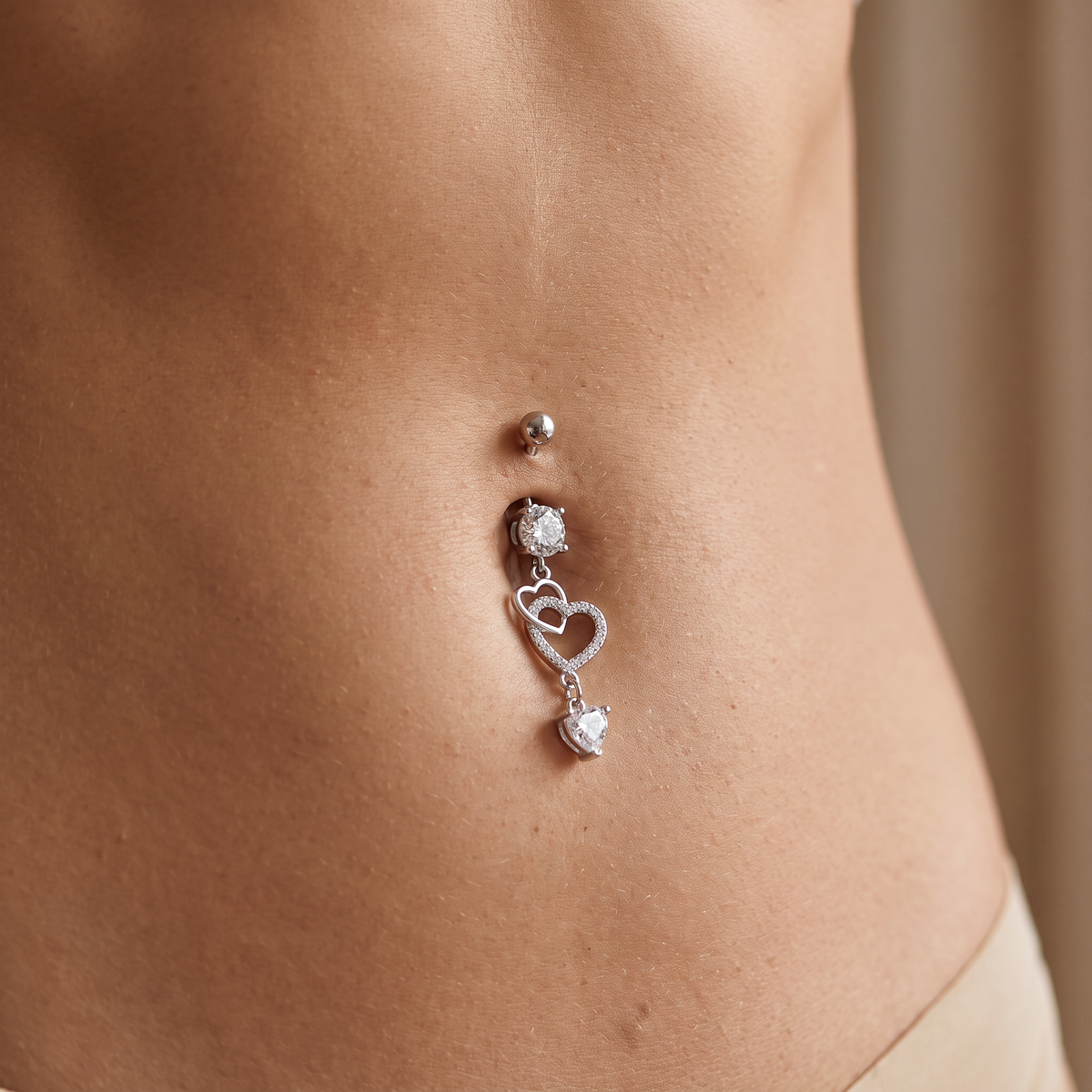 A close-up of a silver belly button ring with a ball stud, a round bezel-set stone, and a dangling double-heart charm with clear stones, shown worn on a person's abdomen.