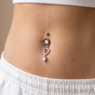 A close-up of a belly button piercing with a silver-toned chain, a small heart-shaped charm, and clear rhinestone accents, worn on a person's midsection.