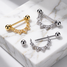 Three curved barbell body jewelry piercings with decorative scalloped front charms and ball end beads are arranged on a white marble slab. Two pieces are gold-toned and one is silver-toned; one matching ball stud is placed beside them.