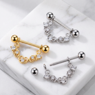 Two curved barbell piercings with decorative scrollwork and clear stones, shown in gold and silver finishes on a white marble surface.
