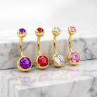 Four gold curved barbell belly button piercings with round faceted gemstones in purple, red, clear, and pink, displayed on a white marble surface.