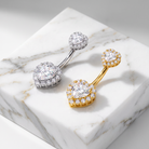 Two curved barbell belly button rings with halo-set round clear stones, one in silver-toned metal and one in yellow-gold metal, displayed on a white marble block.