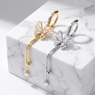 Two butterfly-shaped hoop earrings displayed on a white marble block, one in yellow gold and one in silver, both adorned with clear crystal pavé detailing and dangling crystal drops.