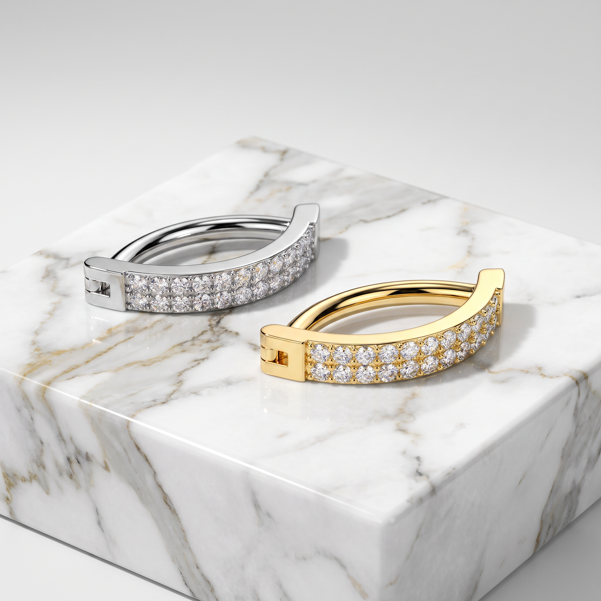 Two curved hoop-style rings with rows of clear stones, one silver-toned and one yellow gold-toned, displayed on a white marble block against a light gray studio background.