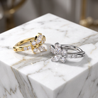 Two flower-shaped rings with marquise-cut clear stones, one in yellow gold and one in silver, displayed on a white marble block.