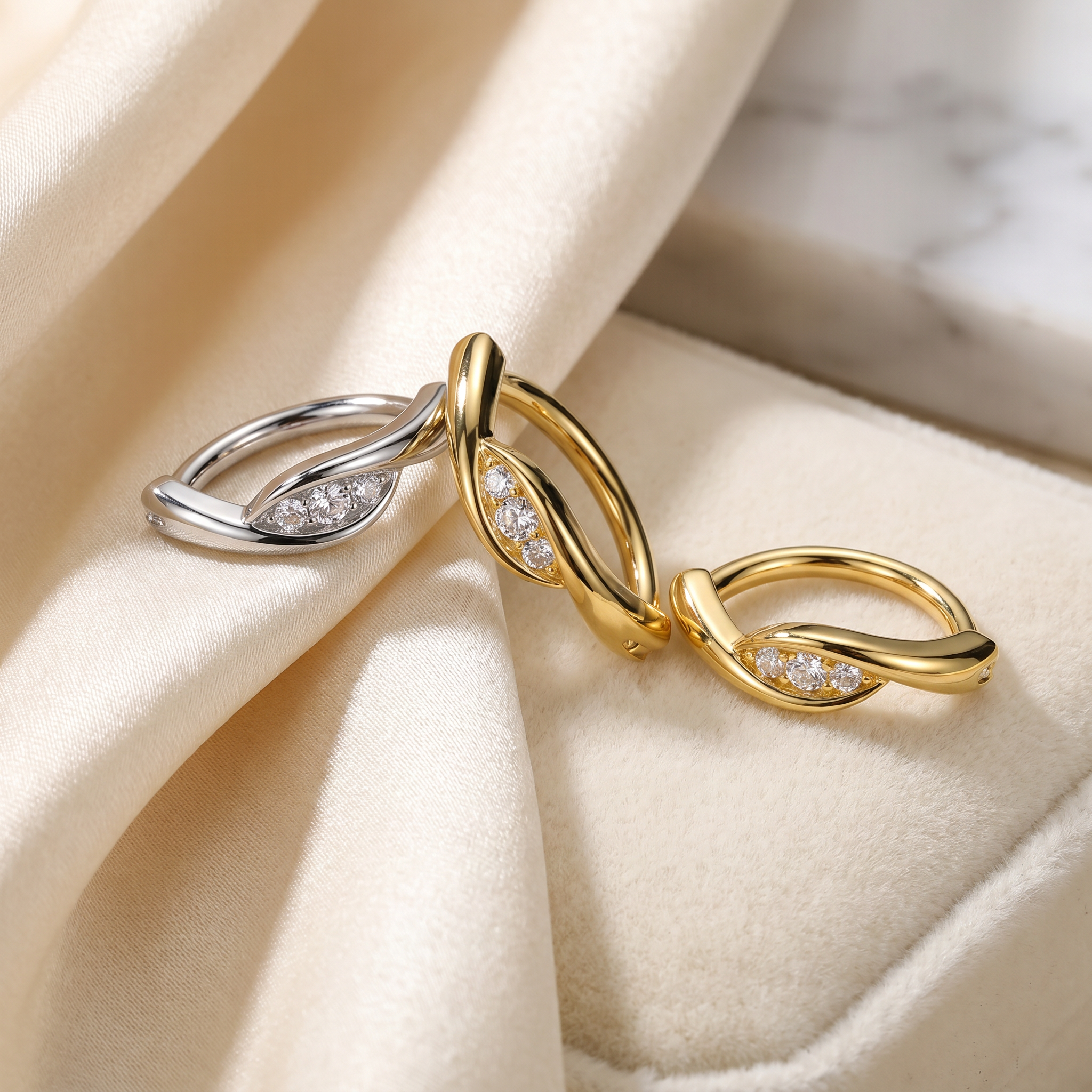 Two twisted leaf-shaped rings with small clear stones, one in yellow gold and one in silver, displayed on a cream satin fabric surface.