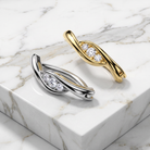 Two twisted leaf-shaped earrings with pavé-set clear stones, shown on a white marble block. One earring is yellow gold and the other is silver, photographed in a clean studio setting.