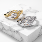 Two elegant twist-style rings with large round center stones and rows of small pavé stones, shown on a white marble display block. One ring is yellow gold with silver-toned accents, and the other is silver-toned with gold-toned accents.