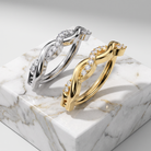 Two twisted hoop-style rings with pavé-set clear stones, shown in silver and yellow gold tones, displayed on a white marble block against a light gray studio background.