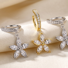 Three flower-shaped hoop earrings with marquise-cut clear stones are displayed on a soft beige fabric background. Two earrings are silver-toned and one is gold-toned, arranged symmetrically on draped satin.