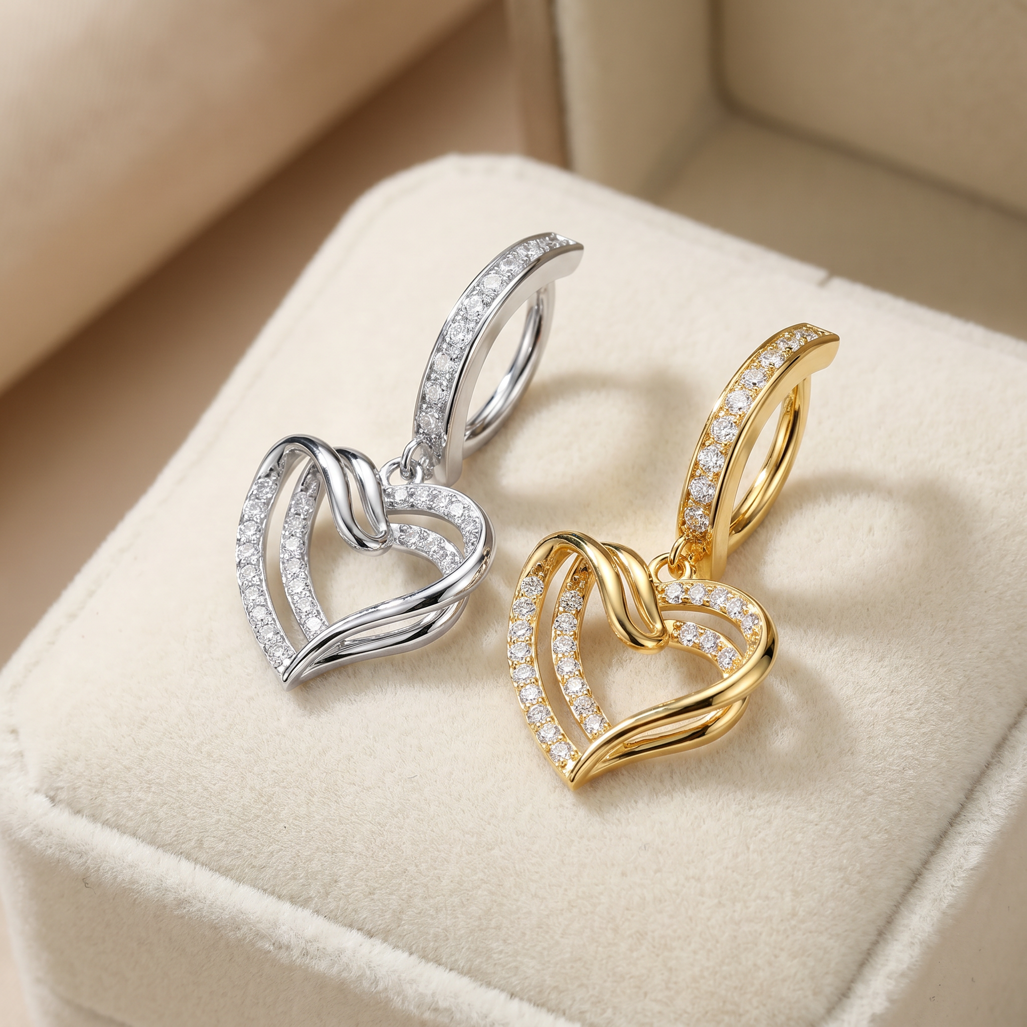 Two double-heart hoop earrings with pavé-set clear stones, shown in silver and gold finishes resting on a cream-colored jewelry box.