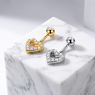 Two heart-shaped barbell belly button piercings displayed on a white marble cube. One is gold with clear rhinestones, and the other is silver with clear rhinestones, set against a light gray background.