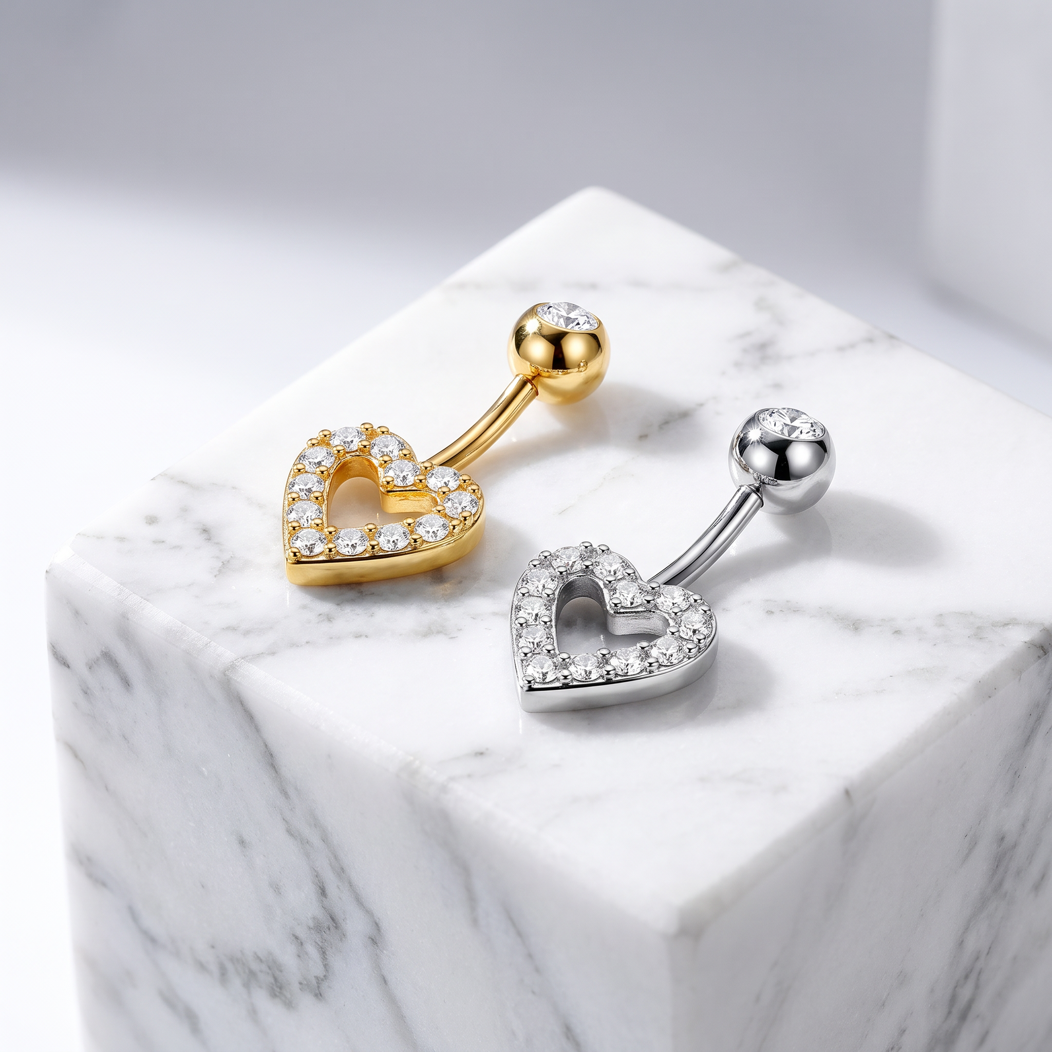 Two heart-shaped barbell belly button piercings displayed on a white marble cube. One is gold with clear rhinestones, and the other is silver with clear rhinestones, set against a light gray background.