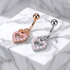 Two belly button rings displayed on a white marble block. One ring is rose gold with a pink heart-shaped crystal charm, and the other is silver with a pink heart-shaped crystal charm.