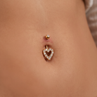 A close-up of a person's lower torso showing a gold belly button ring with a heart-shaped crystal border and a small gold stud above it.