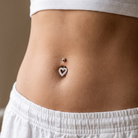 A close-up of a person's midsection wearing a silver belly button ring with a heart-shaped crystal design, paired with a white ribbed crop top and white drawstring shorts.