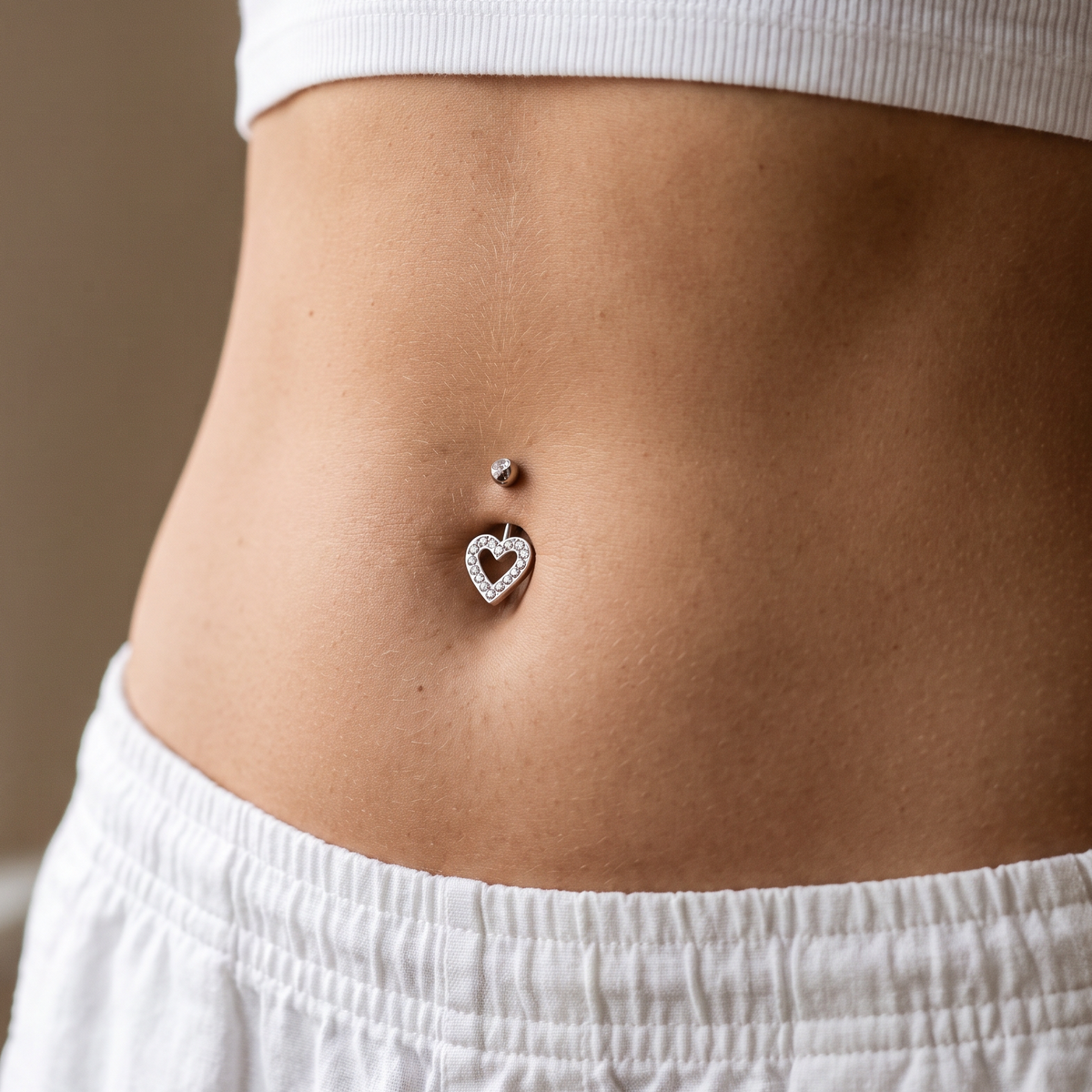 A close-up of a person's midsection wearing a silver belly button ring with a heart-shaped crystal design, paired with a white ribbed crop top and white drawstring shorts.