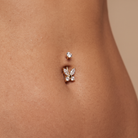 A close-up of a person's lower abdomen showing a rose gold butterfly belly button ring with clear crystals, alongside a smaller matching rose gold stud earring.