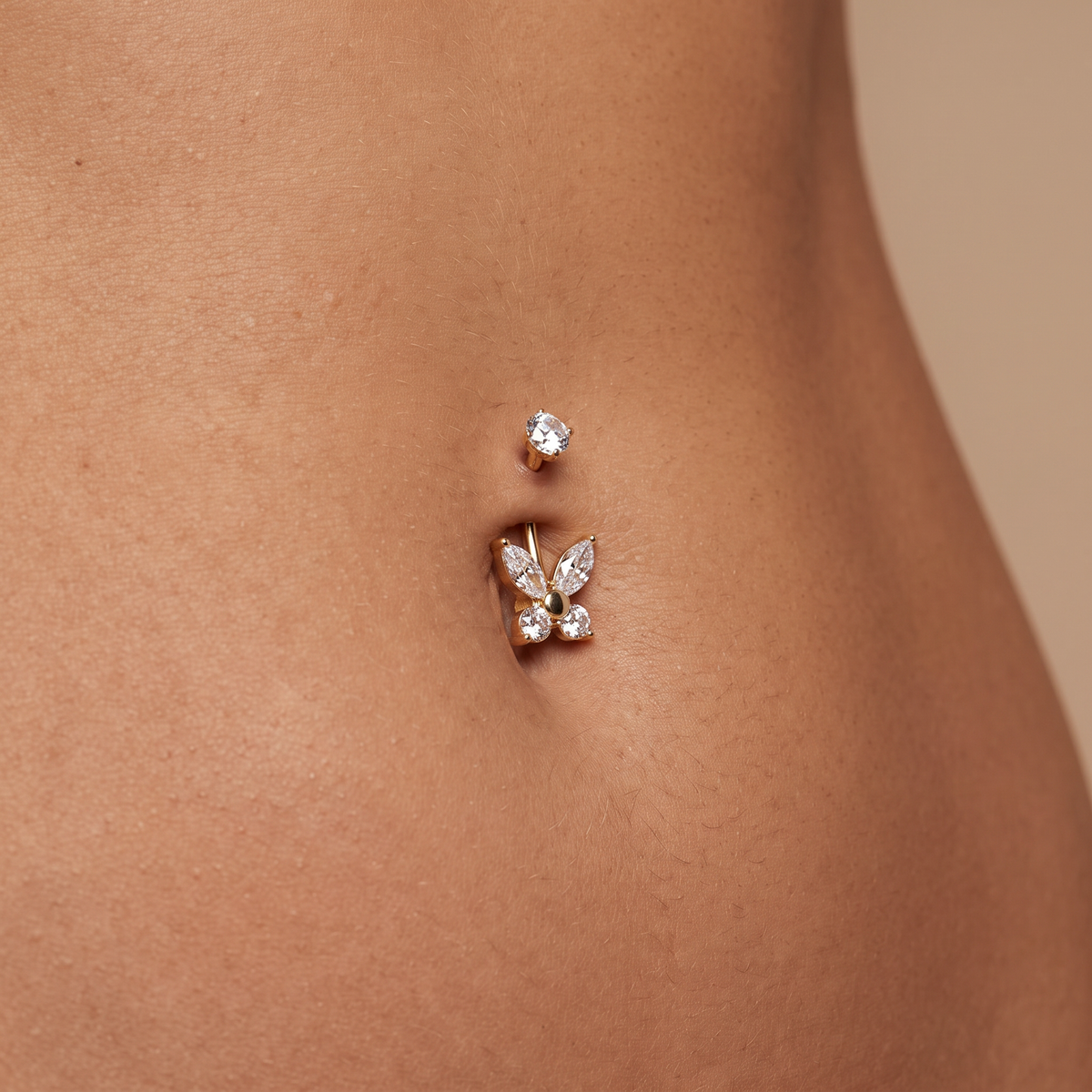 A close-up of a person's lower abdomen showing a rose gold butterfly belly button ring with clear crystals, alongside a smaller matching rose gold stud earring.