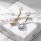 Two curved barbell-style body jewelry pieces with princess-cut clear stones, shown on a white marble block. One piece is gold-toned and the other is silver-toned.