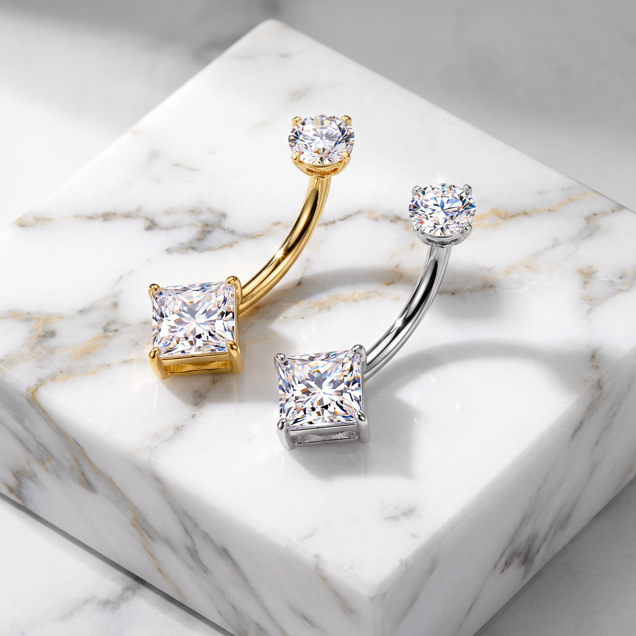 Two curved barbell-style body jewelry pieces with princess-cut clear stones, shown on a white marble block. One piece is gold-toned and the other is silver-toned.