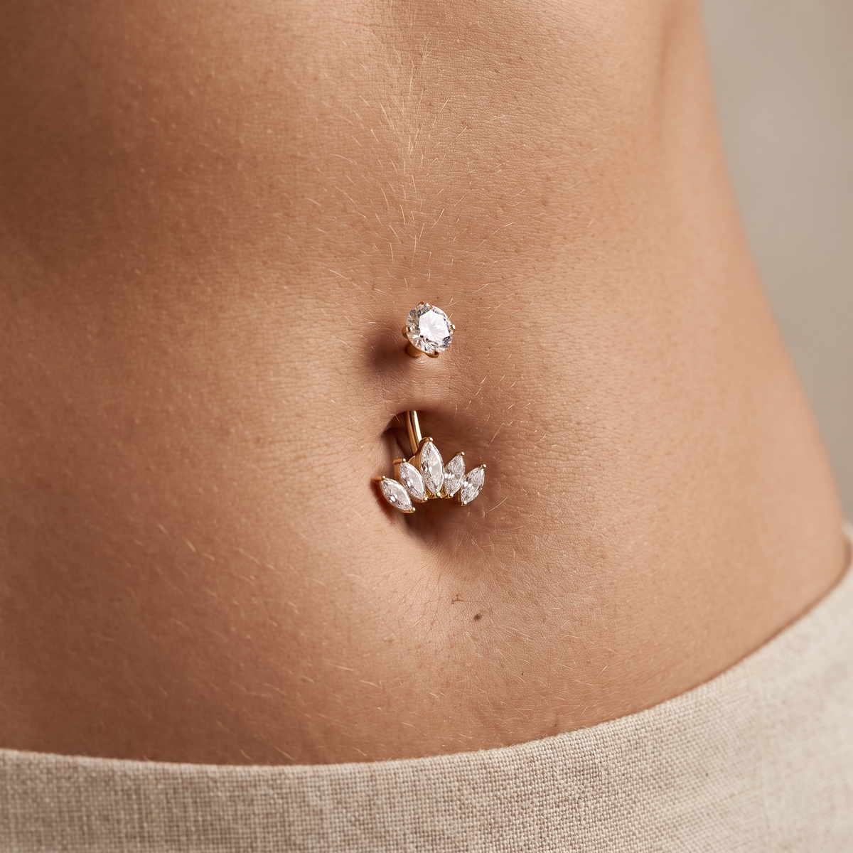 A close-up of a person's lower torso showing a gold belly button ring with a small clear gemstone stud above it and a lotus flower cluster of clear crystals on the main barbell, worn against light beige fabric.