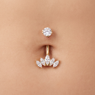 A close-up of a gold navel piercing with a floral leaf cluster setting and a matching stud above it, shown on a person's skin.