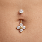 A close-up of a belly button with two gold piercings: a small stud above and a larger belly button ring below featuring a four-petal flower design with clear gemstones and a central gold dot, set against natural skin.