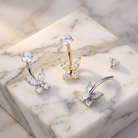 Three butterfly-shaped nose rings with clear crystal gemstones, shown in silver and gold metal finishes, arranged on a white marble display block.