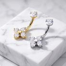 Two flower-shaped belly button rings, one in yellow gold and one in silver, each featuring a round clear crystal in the center and four smaller clear crystals around the petals, displayed on a white marble block.