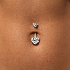 A close-up of a belly button piercing with a gold barbell featuring two heart-shaped clear gemstones, shown on a person's skin.
