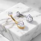 Two heart-shaped stud piercings displayed on a white marble block. One has a yellow gold curved bar and bezel-set heart-shaped clear stone, while the other has a silver-colored curved bar and a pear-cut clear stone.
