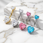 Four heart-shaped barbell belly button piercings arranged diagonally on a white marble slab. The jewelry features heart-shaped and round-cut clear, silver, pink, and blue stones in gold, silver, and white metal settings.