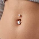 A close-up of a person's navel area showing a gold belly button ring with a heart-shaped clear gemstone and a smaller matching stud above it.