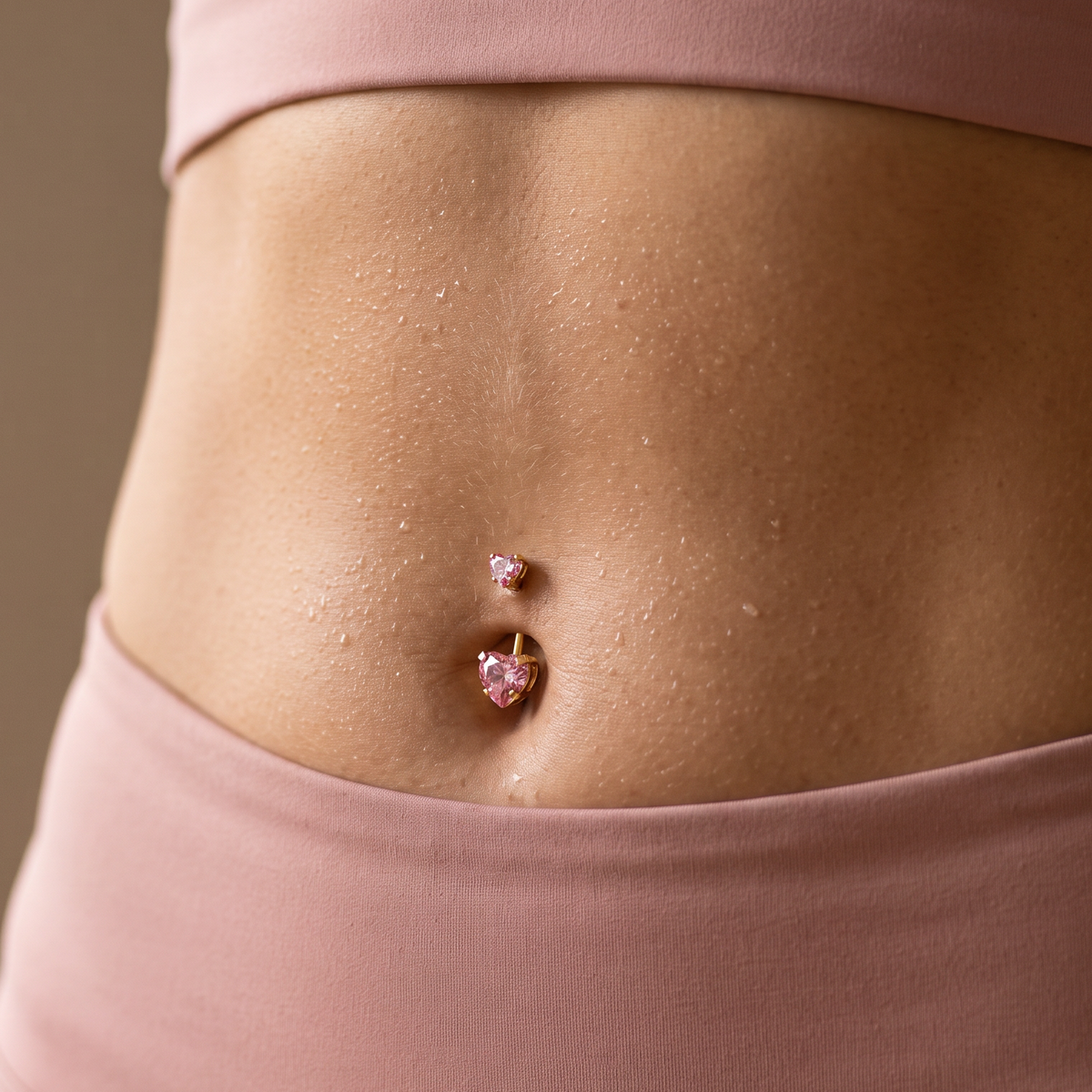 A close-up of a person's midsection wearing a dusty pink sports bra and high-waisted leggings, featuring a pink heart-shaped stud belly button piercing.