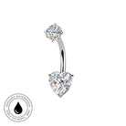 A curved barbell belly button ring with a heart-shaped clear gemstone on the bottom and a round clear gemstone on the top, set in silver-toned metal. A circular badge in the bottom-left corner reads 'SKIN SAFE WATERPROOF AND TARNISH FREE' with a black droplet symbol.