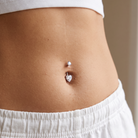 A close-up of a person's midsection showing a belly button piercing with two silver-toned barbells and a small heart-shaped charm.