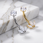 Two curved barbell belly button piercings with marquise-cut clear stones, displayed on a white marble block and marble surface. One is silver-toned and the other is gold-toned.