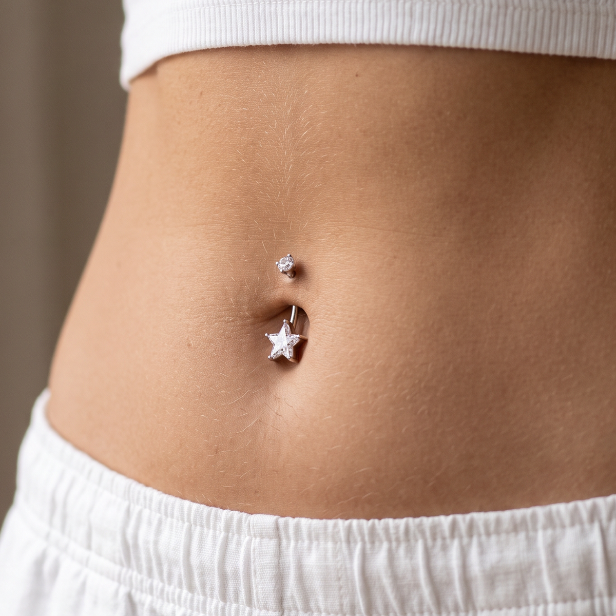 A close-up of a person's midsection wearing a silver belly button ring with a small star charm and a tiny stud, paired with a white ribbed crop top and white shorts.