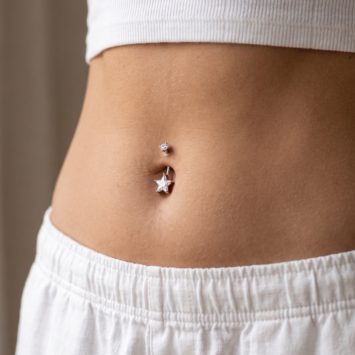 A close-up of a person's midsection wearing a silver belly button ring with a small star charm and a tiny stud, paired with a white ribbed crop top and white shorts.