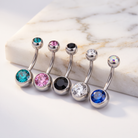 Five silver-toned belly button rings with round faceted gemstones in turquoise, pink, black, clear, and blue, arranged on a white marble slab against a white background.